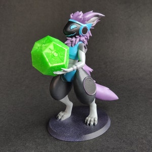 Puede incluir: Una figura peluda de color morado y negro con un objeto geométrico verde en la mano. La figura lleva un atuendo azul y blanco y tiene un auricular blanco y negro en la cabeza. La figura está de pie sobre una base morada.