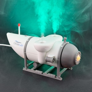 May include: A white and gray submarine-shaped diffuser with the OceanGate logo. The diffuser has a red antenna and a light emanating from the front. It sits on a gray stand, with green vapor rising from the top.