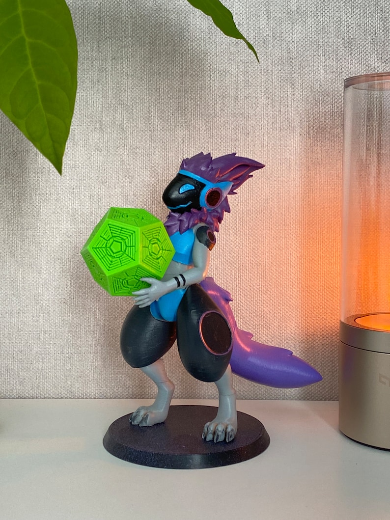 May include: A 3D printed figurine of a purple and blue creature holding a green geometric object. The creature has a black face mask and a blue and white outfit. The figurine is standing on a black base.