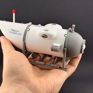 K&ouml;nnte beinhalten: Ein Modell-U-Boot in Wei&szlig; und Grau, gehalten in einer Hand. Das Modell hat das "OceanGate"-Logo und eine rote Antenne. Das U-Boot ruht auf einer grauen St&uuml;tzstruktur.