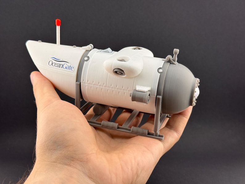 K&ouml;nnte beinhalten: Ein Modell-U-Boot in Wei&szlig; und Grau, gehalten in einer Hand. Das Modell hat das "OceanGate"-Logo und eine rote Antenne. Das U-Boot ruht auf einer grauen St&uuml;tzstruktur.