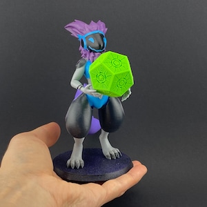 May include: A 3D printed figurine of a purple and blue furry creature holding a green geometric object. The figurine is standing on a black base.