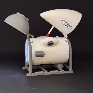 K&ouml;nnte beinhalten: Ein 3D-gedrucktes Modell eines zylindrischen U-Boots mit dem Text "OceanGate". Das Modell ist haupts&auml;chlich wei&szlig; und grau, mit einem roten Akzent. Es verf&uuml;gt &uuml;ber eine offene Luke und ist auf einer grauen rechteckigen Basis montiert. Das Modell &auml;hnelt einem U-Boot.