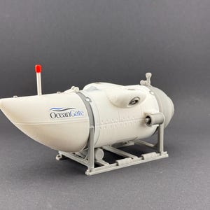 K&ouml;nnte beinhalten: Ein Modell eines wei&szlig;en und grauen U-Boots mit dem Text "OceanGate" an der Seite. Das Modell hat eine rote Antenne und ist auf einem grauen St&auml;nder montiert. Das U-Boot hat eine zylindrische Form.