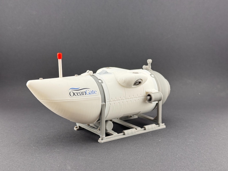 K&ouml;nnte beinhalten: Ein Modell eines wei&szlig;en und grauen U-Boots mit dem Text "OceanGate" an der Seite. Das Modell hat eine rote Antenne und ist auf einem grauen St&auml;nder montiert. Das U-Boot hat eine zylindrische Form.