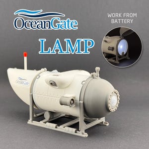 K&ouml;nnte beinhalten: Eine wei&szlig;-graue Lampe in Form eines U-Boots mit dem blauen Text "OceanGate LAMP". Die Lampe hat eine rote Antenne und ein kleines eingebettetes Bild, das die beleuchtete Lampe zeigt. Der Text "WORK FROM BATTERY" ist ebenfalls sichtbar.