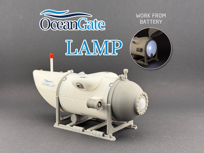 K&ouml;nnte beinhalten: Eine wei&szlig;-graue Lampe in Form eines U-Boots mit dem blauen Text "OceanGate LAMP". Die Lampe hat eine rote Antenne und ein kleines eingebettetes Bild, das die beleuchtete Lampe zeigt. Der Text "WORK FROM BATTERY" ist ebenfalls sichtbar.