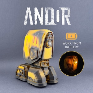 May include: A yellow and black robot-like lamp with the word "ANDOR" above it. The lamp has a circular light on the front and the text "WORK FROM BATTERY". The lamp is on a gray background.