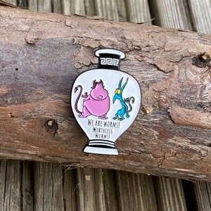 May include: White enamel pin featuring two cartoon characters inside a white vase with a black outline. The characters are a pink monster with horns and a blue monster with horns. The text on the pin reads "We are worms! Worthless worms!"