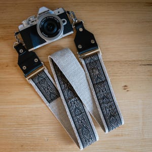 Könnte beinhalten: Ein Kameragurt aus beigem Webstoff mit einem schwarzen Zierornament. Der Gurt hat schwarze Lederenden und Metallbeschläge. Eine Olympus OM-D Kamera ist ebenfalls im Bild zu sehen.