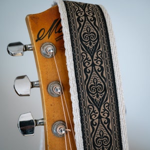 May include: A close-up shot of a guitar headstock with a patterned guitar strap. The strap has a black and tan design with a cream border. The wooden headstock has silver tuning pegs. The guitar is vintage.
