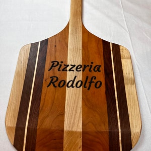 May include: A wooden pizza peel with a long handle. The peel is made of multiple types of wood, including cherry, maple, and walnut. The peel is engraved with the text "Pizzeria Rodolfo".
