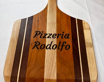 May include: A wooden pizza peel with a long handle. The peel is made of multiple types of wood, including cherry, maple, and walnut. The peel is engraved with the text "Pizzeria Rodolfo".