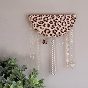 May include: A half-circle jewelry organizer with a leopard print design. Several necklaces, earrings, and a ring hang from the organizer. The organizer is mounted on a gray wall. Green plants in white pots are in the foreground.