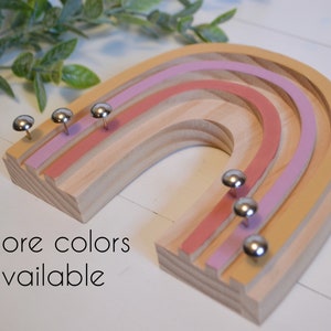 Wood rainbow shape with arches painted in retro colors of deep orange, mauve and mustard yellow. The wood between the arches is a light wood color. There are six shiny silver tacks near the bottom of the rainbow to hang necklaces.