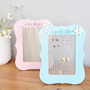 May include: Two light blue and pink earring holders with white flower accents. The holders have a grid pattern and the names "evelyn" and "natalie" are written on them.