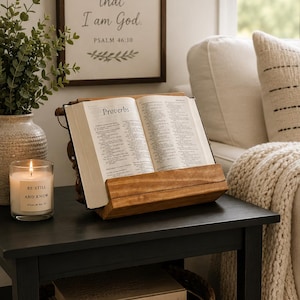 May include: A wooden book stand holding an open Bible to the book of Proverbs. The stand is placed on a black side table next to a lit candle in a glass jar with a label reading Be Still and Know Psalm 46:10, and a woven basket containing two books.