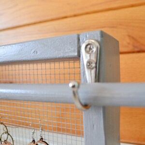 May include: A gray wooden earring display with a wire mesh backing and a horizontal rod. The display is accented with silver hardware and is designed to hold various styles of earrings. The background is a wooden wall.