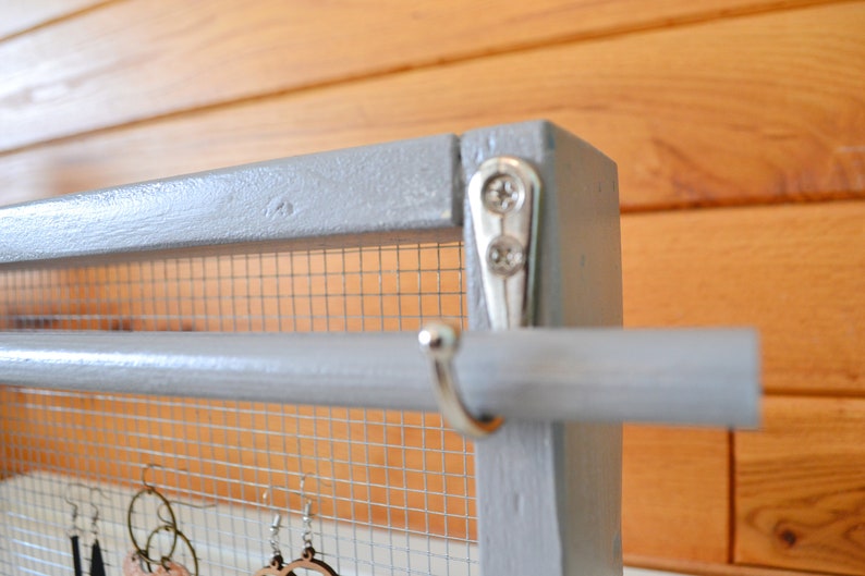 May include: A gray wooden earring display with a wire mesh backing and a horizontal rod. The display is accented with silver hardware and is designed to hold various styles of earrings. The background is a wooden wall.
