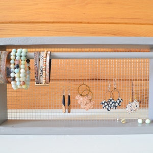 May include: A gray wooden jewelry organizer with a mesh backing. The organizer displays various bracelets and earrings. Bracelets include beaded and leather styles. Earrings include hoop and drop styles in various colors and patterns.