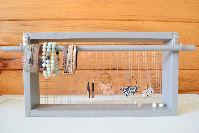 May include: A gray wooden jewelry organizer with a mesh backing. The organizer displays various bracelets and earrings. Bracelets include beaded and leather styles. Earrings include hoop and drop styles in various colors and patterns.