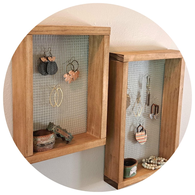 This is a wood wall hanging earring holder. The frame is rectangular and the wood is stained with a medium brown color. There is square chicken wire on the backing to hold dangle earrings. The style is simple, minimalistic and a little rustic.