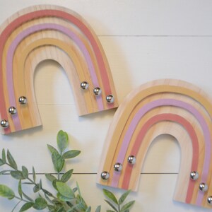 May include: Two wooden rainbow-shaped wall decorations. Each rainbow has layers of light wood, pink, and yellow. Small silver knobs are attached to the bottom of each rainbow. The background is a white wooden surface.