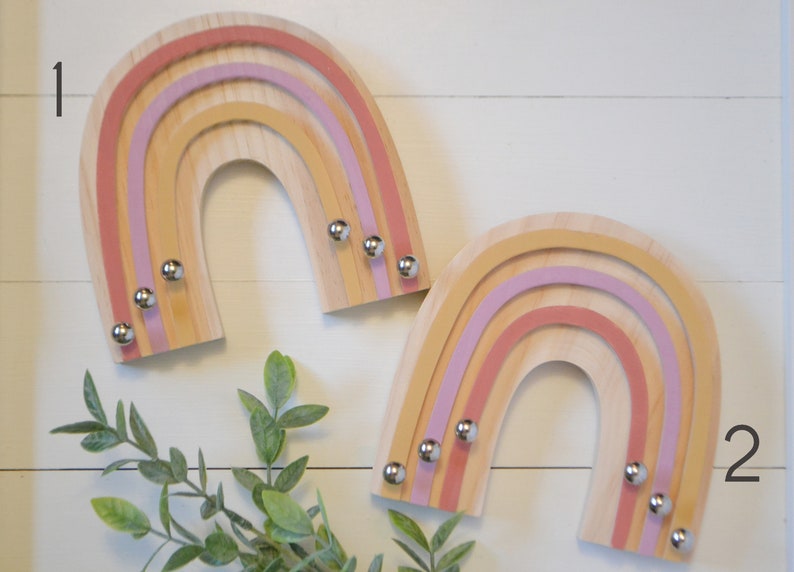 May include: Two wooden rainbow-shaped wall decorations. Each rainbow has layers of light wood, pink, and yellow. Small silver knobs are attached to the bottom of each rainbow. The background is a white wooden surface.