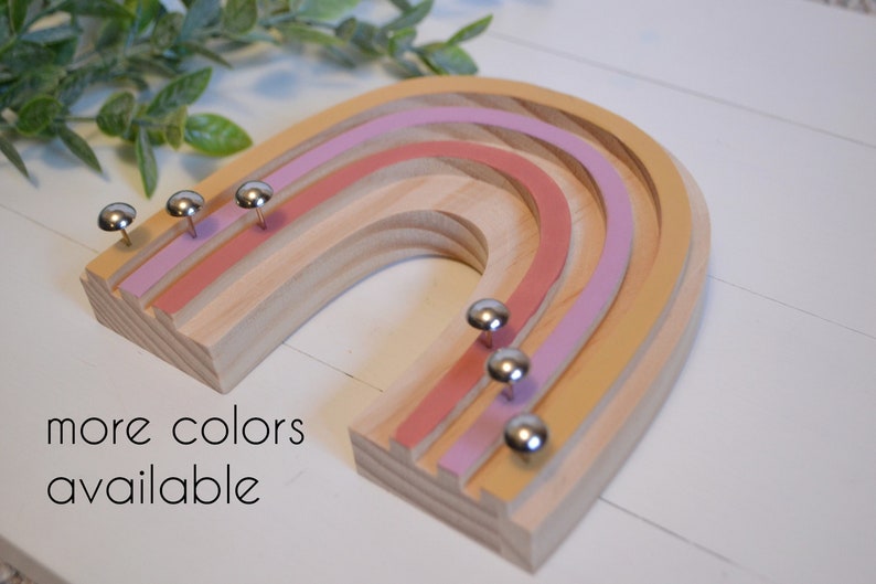 Wood rainbow shape with arches painted in retro colors of deep orange, mauve and mustard yellow. The wood between the arches is a light wood color. There are six shiny silver tacks near the bottom of the rainbow to hang necklaces.