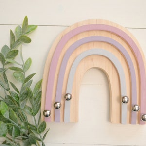 May include: A wooden rainbow wall decor with three layers of pastel pink, purple, and gray paint. The rainbow has four silver metal hooks.