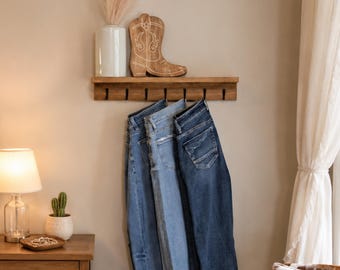 May include: A wooden wall-mounted rack with six black metal hooks, holding three pairs of denim jeans in varying shades of blue. The rack is positioned below a white vase and a wooden cowboy boot cutout decoration on a shelf.