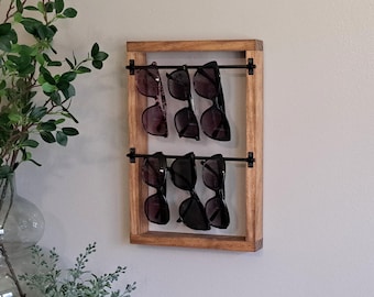 May include: A rectangular wooden frame with a medium brown finish featuring two horizontal black metal rods. Six pairs of dark-tinted sunglasses are hung from the rods, with three pairs suspended from the top bar and three from the bottom bar.