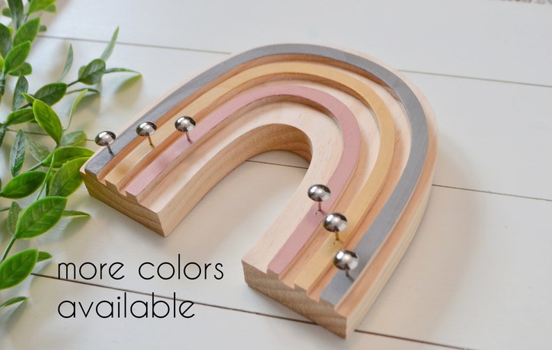 Wood rainbow shape with arches painted in muted shades of pink, yellow and blue gray. The wood between the arches is a light wood color. There are six shiny silver tacks near the bottom of the rainbow to hang necklaces.