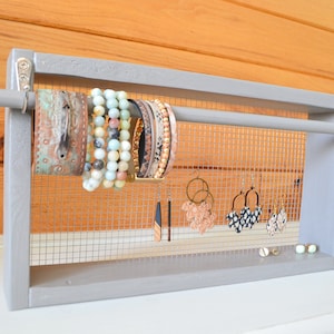 May include: A gray wooden jewelry organizer with a mesh backing. The organizer holds various bracelets and earrings. Bracelets include beaded and leather styles. Earrings include hoop and dangle designs. The background is a light wood.