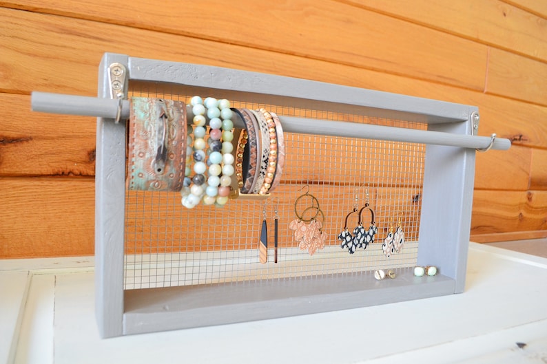 May include: A gray wooden jewelry organizer with a mesh backing. The organizer holds various bracelets and earrings. Bracelets include beaded and leather styles. Earrings include hoop and dangle designs. The background is a light wood.