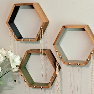 May include: Three wooden hexagon shelves with a natural wood finish are mounted on a light gray wall. Each shelf has a dark interior and small silver knobs along the bottom edge. Gold necklaces hang from the shelves.