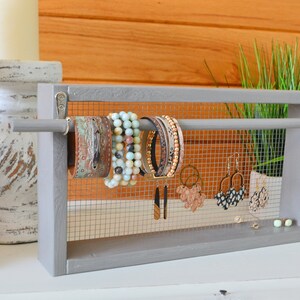 May include: A gray wooden jewelry organizer with a wire mesh backing. The organizer holds various bracelets and earrings. The bracelets are leather, beaded, and multi-strand. Earrings include gold hoops and patterned studs. The background features candles and greenery.