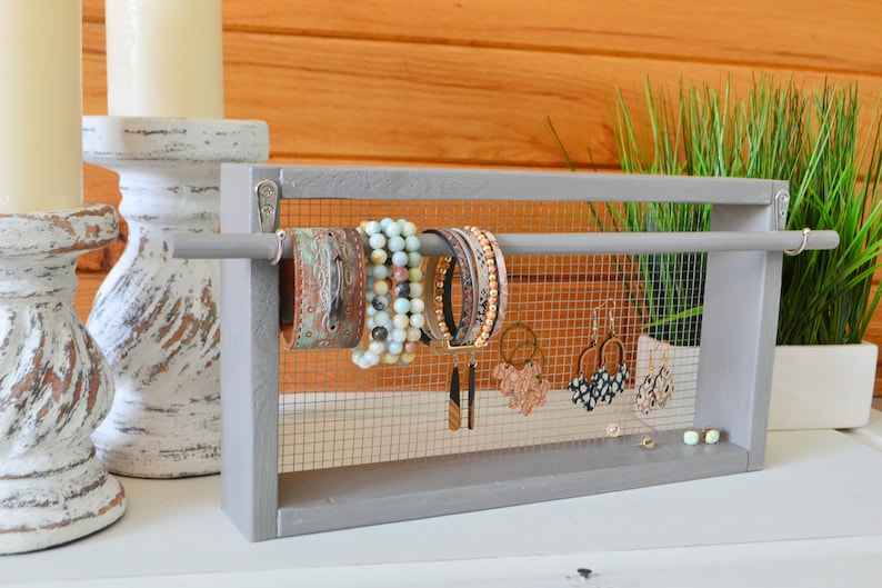 May include: A gray wooden jewelry organizer with a wire mesh backing. The organizer holds various bracelets and earrings. The bracelets are leather, beaded, and multi-strand. Earrings include gold hoops and patterned studs. The background features candles and greenery.