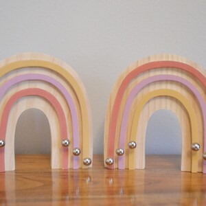 May include: Two wooden rainbow-shaped decorative objects. Each rainbow has layers of pastel colors: yellow, pink, and purple. Small silver spheres are attached to the bottom of each rainbow. The rainbows are displayed on a wooden surface.