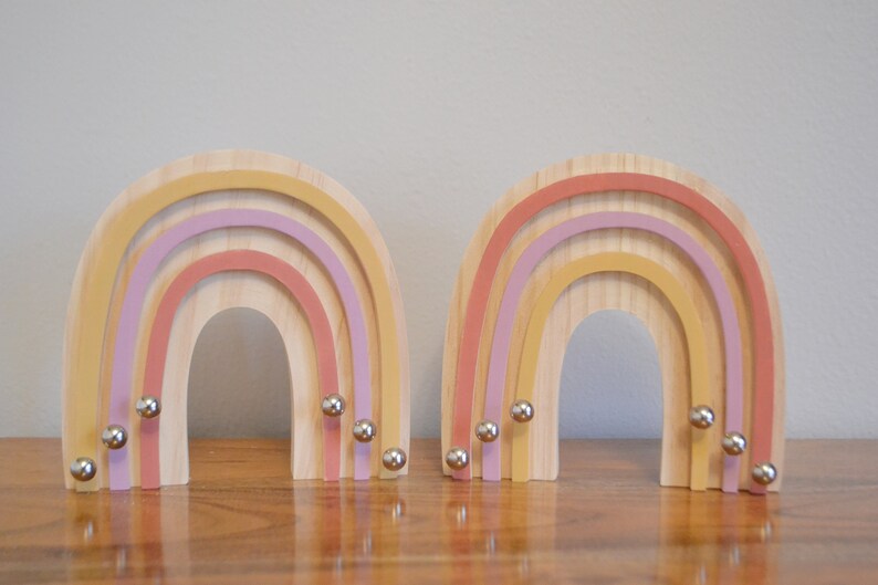 May include: Two wooden rainbow-shaped decorative objects. Each rainbow has layers of pastel colors: yellow, pink, and purple. Small silver spheres are attached to the bottom of each rainbow. The rainbows are displayed on a wooden surface.