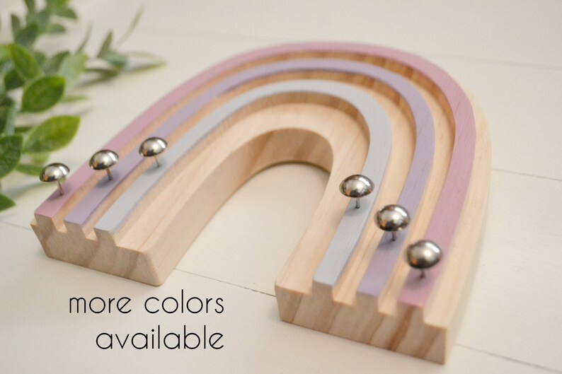 Wood rainbow shape with arches painted in shades of purple and gray. The wood between the arches is a light wood color. There are six shiny silver tacks near the bottom of the rainbow to hang necklaces.