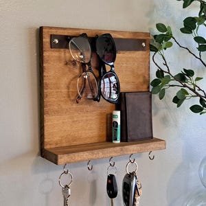 May include: A wall-mounted wooden organizer featuring a horizontal leather strap for hanging two pairs of glasses, a small shelf holding a brown wallet and a tube of lip balm, and five metal hooks underneath for keys and other small hanging items.