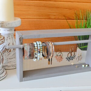 May include: A gray wooden jewelry organizer with a mesh backing, displaying bracelets and earrings. The organizer has a rod for hanging jewelry and is placed on a white surface. Two white candles in distressed white candle holders are in the background.