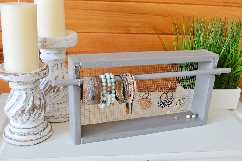 May include: A gray wooden jewelry organizer with a mesh backing, displaying bracelets and earrings. The organizer has a rod for hanging jewelry and is placed on a white surface. Two white candles in distressed white candle holders are in the background.