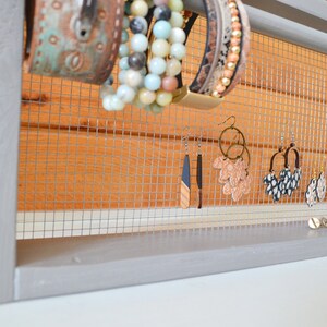 May include: A gray wooden jewelry organizer with a wire mesh backing. The organizer displays various earrings and bracelets. The bracelets are in shades of turquoise, beige, and brown. The earrings are in various styles and colors.