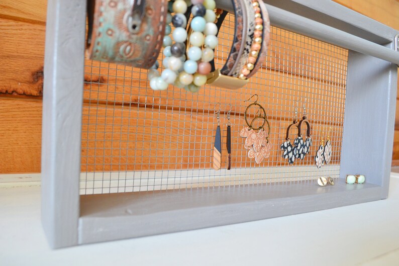 May include: A gray wooden jewelry organizer with a wire mesh backing. The organizer displays various earrings and bracelets. The bracelets are in shades of turquoise, beige, and brown. The earrings are in various styles and colors.