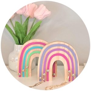 May include: Two wooden rainbow decorations with colorful stripes in pink, blue, green, and purple. Each rainbow has silver ball accents and a gold chain. A vase of pink tulips is in the background.