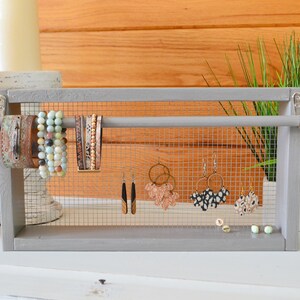 May include: A gray wooden jewelry organizer with a wire mesh backing. The organizer displays various bracelets and earrings. The bracelets are in shades of brown, turquoise, and gold. The earrings include gold hoops and patterned designs.