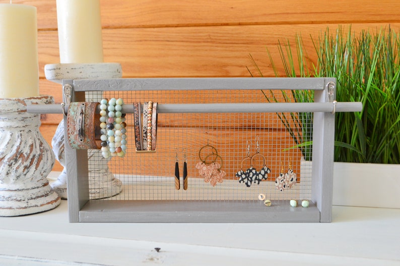 May include: A gray wooden jewelry organizer with a wire mesh backing. The organizer displays various bracelets and earrings. The bracelets are in shades of brown, turquoise, and gold. The earrings include gold hoops and patterned designs.