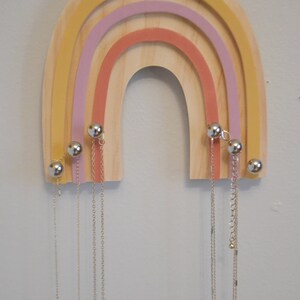 May include: Wooden rainbow-shaped jewelry organizer with a natural wood finish and colorful arches in yellow, pink, and coral. Silver-colored ball-shaped knobs hold multiple necklaces of varying lengths. A decorative and functional item.